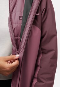 Paarse geïsoleerde jas met ritsdetail en logo, gemaakt van soepele, structuurstof. Hand die de rits vasthoudt, toont de binnenvoering.