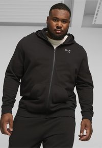 Sort zip-up hoodie lavet af blødt stof, med en kengurulomme, hætte og hvidt logo-accents på brystet.