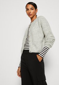 Light grey, knit cardigan with a round neckline, front buttons, and ribbed cuffs. Worn over a black and white striped top and black pants.