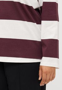 Une personne portant une chemise à manches longues avec des rayures horizontales bordeaux et blanches, partiellement visible sur un fond sombre.