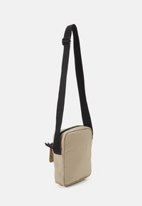 Borsa a tracolla in tessuto beige con forma rettangolare, tracolla nera regolabile, chiusura con zip e una piccola tasca frontale.