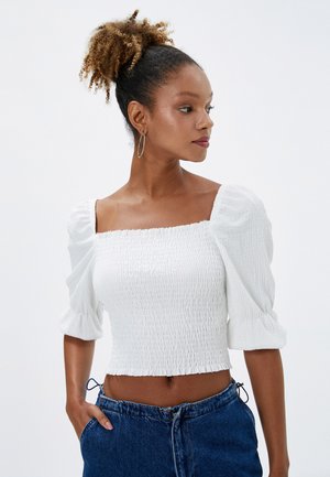 Vit smockad kort topp med fyrkantig halsringning och puffärmar. I kombination med en högmidjad denimkjol som visar en avslappnad, texturerad stil.