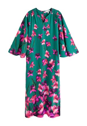 Kvinde med langt mørkt hår iført grøn bluse med lyserødt blomsterprint og gyldne dråbeformede øreringe, smilende let og kigger mod venstre.