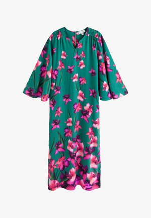 Kvinde med langt mørkt hår iført grøn bluse med lyserødt blomsterprint og gyldne dråbeformede øreringe, smilende let og kigger mod venstre.