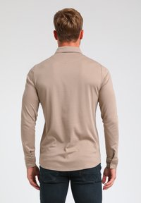 Beige Polohemd mit langen Ärmeln, kragenlosem Hals, glatter Textur und geknöpften Manschetten. Trägt dazu eine dunkle Jeanshose mit geschwungenem Saum.