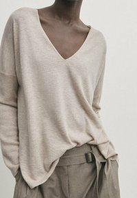 Pull en tricot beige à col en V avec une coupe décontractée, manches longues, texture douce et une finition côtelée discrète, associé à un pantalon taille haute gris.