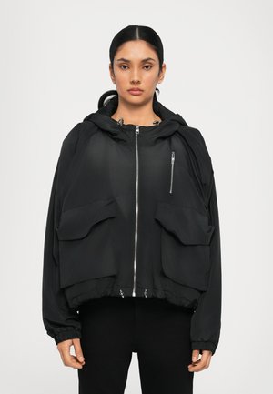 Sort cropped jakke med hætte, der har en frontlynlås, to store klaplommer og elastiske manchetter. Glat tekstur, letvægtigt materiale.