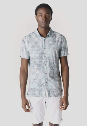 Joven de pie, vestido con una camisa de manga corta estampada de color azul claro y pantalones cortos blancos arremangados, sobre un fondo neutro.