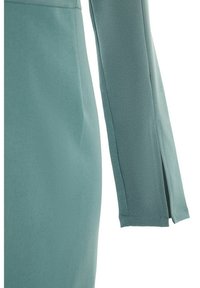 Détail de la manche et du côté d’un vêtement ajusté bleu sarcelle avec une couture verticale et une petite fente au poignet.