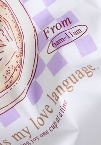 Zbliżenie na tkaninę z ilustracją bajgla, fioletowe tło w kratę oraz tekst odwołujący się do "Od 6 rano do 11 rano" i "mój język miłości."