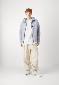 Felpa grigia chiara con dettagli neri, t-shirt bianca, pantaloni cargo beige e sneaker alte beige. Tessuto morbido, vestibilità rilassata.