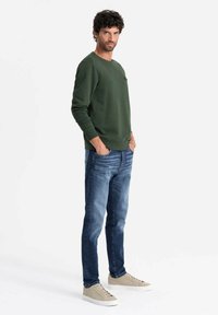 Felpa verde con scollatura a girocollo, vestibilità casual. Jeans azzurri chiari, con gambe taper. Sneakers beige con suola piatta e design con lacci.