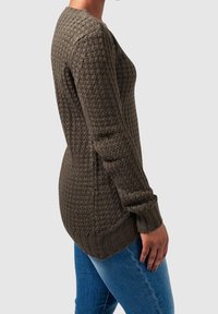 Femme portant un pull en maille vert olive foncé avec un motif texturé et un jean bleu, se tenant de profil sur un fond neutre.