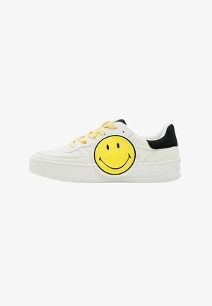White sneaker with a round yellow smiley face patch on the side, black heel accent, and yellow laces, featuring a textured rubber sole.