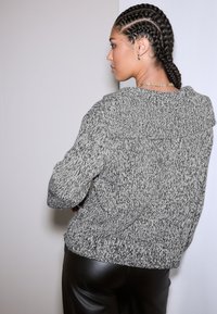 Sivi pleteni džemper s teksturiranim uzorkom, opuštenog kroja i ovratnikom poput sal ovratnika, upareni s crnim kožnim hlačama. Pogled sa stražnje strane prikazuje detalje.