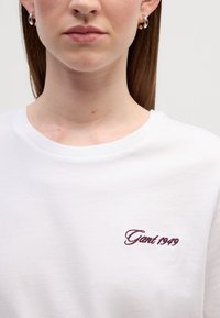 Biała bawełniana koszulka z okrągłym dekoltem. Czerwony haftowany logo "Gant 1949" po lewej stronie klatki piersiowej. Gładka tekstura, casualowy design.