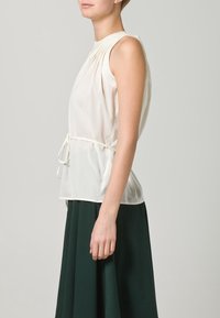 Vit ärmlös topp med veckad detalj vid axeln och midjeband. Slät textur, halvt genomskinligt tyg, kombinerad med en mörkgrön kjol.