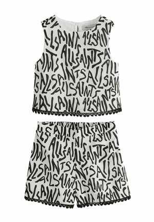 Junges Mädchen, das ein schwarz-weißes ärmelloses Oberteil und passende Shorts mit Textdruck trägt, schwarze Sandalen mit Schnallen auf grauem Hintergrund.