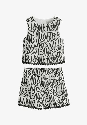 Junges Mädchen, das ein schwarz-weißes ärmelloses Oberteil und passende Shorts mit Textdruck trägt, schwarze Sandalen mit Schnallen auf grauem Hintergrund.