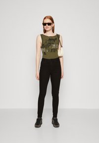 Ελαιογραφίς ριχτό τοπ με ραβδώσεις και γραφική εκτύπωση, συνδυασμένο με μαύρες ψηλόμεσες τζιν παντελόνες και μαύρα αθλητικά παπούτσια. Αξεσουάρ με μια μικρή λευκή τσάντα.