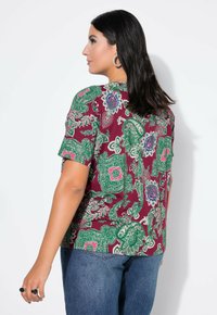 Kurzes Oberteil in Bordeauxrot mit grünem Paisleymuster. Das Material scheint leicht und locker geschnitten zu sein, mit einem runden Halsausschnitt, kombiniert mit blauen Jeans.