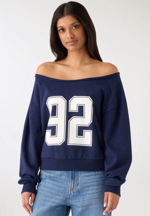 Felpa blu navy con spalle scoperte, grafica bianca "92", bordi a costine, maniche lunghe e vestibilità ampia. Abbinata a jeans di denim chiaro.