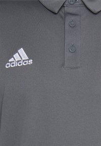 Grijs poloshirt gemaakt van gestructureerde stof. Heeft een drieknoopsluiting en een wit geborduurd Adidas-logo op de linkerborst.