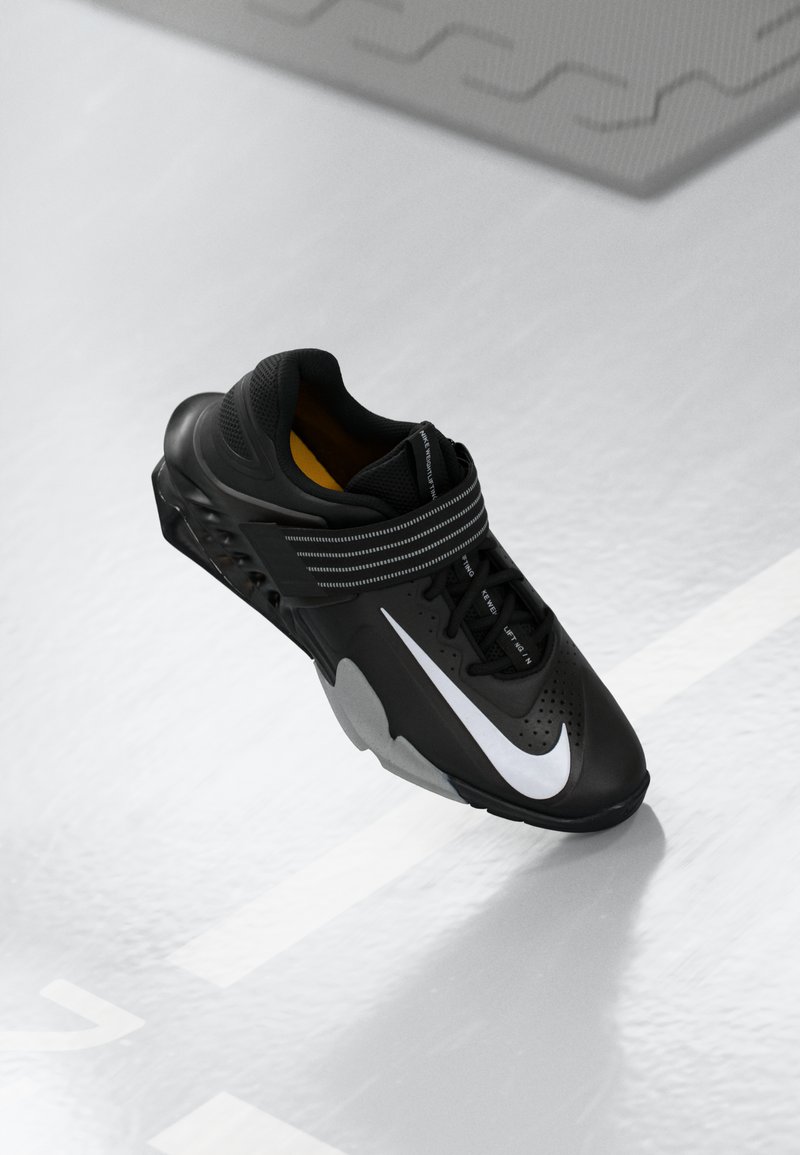 Chaussure de sport noire avec tige en mesh, une virgule blanche Nike bien visible et des accents gris. Présente des sangles et une semelle texturée pour une bonne adhérence.