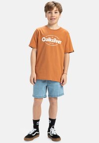 Oranžové bavlnené tričko s bielym logom "Quiksilver", skombinované s svetlomodrými džínsovými šortkami a čiernymi teniskami s bielymi akcentmi a čiernymi ponožkami.