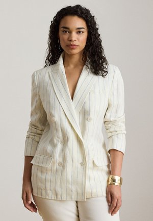 Femme aux cheveux bouclés portant un blazer croisé à fines rayures blanches et un pantalon assorti, avec un bracelet manchette en or à son poignet.