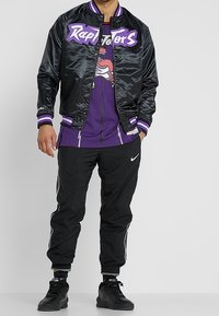 Chaqueta bomber de satén negra con letras moradas, una camiseta de malla morada debajo y pantalones de chándal negros con acentos blancos y el logo de Nike.