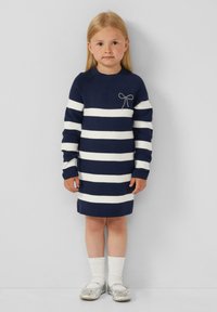 Robe tricot rayée en bleu marine et blanc avec des manches longues, ornée d'un accent en nœud argenté. Associée à des chaussettes blanches montantes et des chaussures argentées.