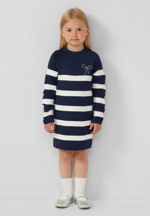 Gestreiftes Strickkleid in Marineblau und Weiß mit langen Ärmeln, versehen mit einem silbernen Schleifenakzent. Kombiniert mit knielangen weißen Socken und silbernen Schuhen.