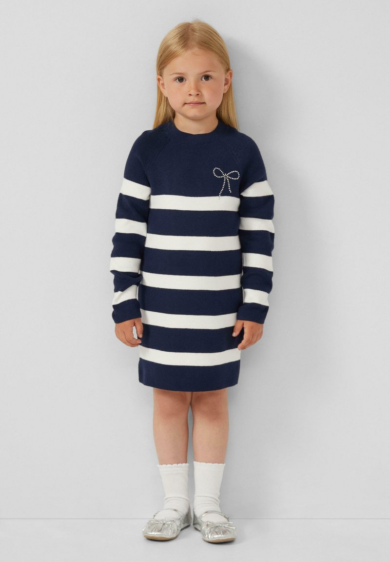 Robe tricot rayée en bleu marine et blanc avec des manches longues, ornée d'un accent en nœud argenté. Associée à des chaussettes blanches montantes et des chaussures argentées.