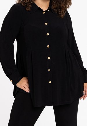 Femme portant une chemise noire à manches longues avec des boutons dorés, debout avec une main sur la hanche.