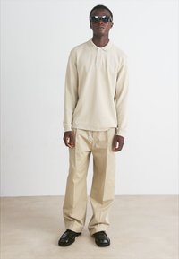 Polo de manga longa bege com colarinho, combinado com calças largas em bege claro e sapatos pretos. Acessórios incluem óculos de sol.