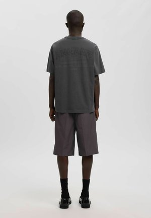 Homme debout de dos portant un t-shirt graphique gris foncé, un short ample foncé, des chaussettes noires et des chaussures habillées noires sur un fond clair uni.