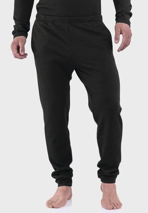 Mann steht barfuß und trägt ein schwarzes Langarmshirt und schwarze enganliegende Jogginghose vor einem schlichten hellen Hintergrund.