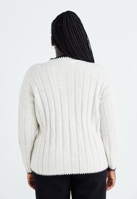 Cream ribbed sweater with long sleeves, featuring a round neckline and a contrasting black scalloped trim along the bottom edge.