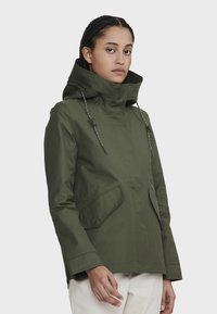 Veste impermeable vert olive avec col haut, capuche ajustable et deux poches latérales. Fabriquée en tissu lisse et durable avec des accents à cordon.