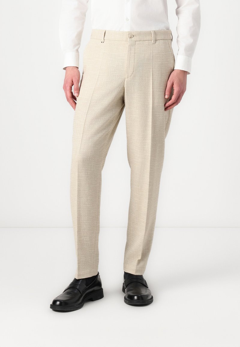 Pantalon texturé beige à coupe droite, avec fermeture par bouton et poches latérales, associé à des mocassins en cuir noirs.
