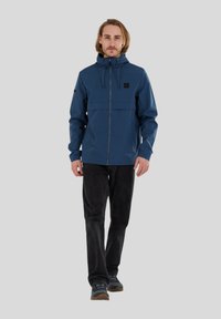 Chaqueta impermeable azul con cremallera frontal, capucha ajustable y dos bolsillos en el pecho. Combinada con pantalones negros y zapatos grises.