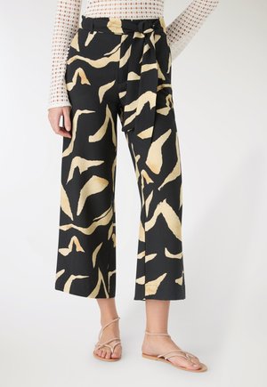 Donna che indossa pantaloni neri a gamba larga e taglio corto con stampa astratta beige e cintura annodata, abbinati a sandali bassi con cinturini.