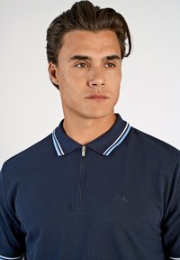 Mörkblå pikétröja med dragkedja i kragen, med ljusblå ränder, texturerat tyg och en subtil logotyp på bröstet.