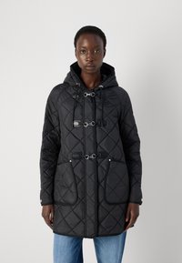 Svart quiltet parka med hette, med sølvfargede lukkinger, to store frontlommer, og en strukturert stofffinish.