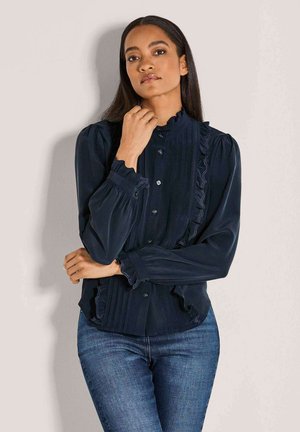 Marineblouse met een gerimpelde kraag en voorkant, lange mouwen met gathered cuffs, knoopsluiting en verticale plooien. Draag met blauwe jeans.
