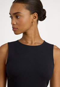 Melna topi bez piedurknēm ar gludu faktūru un apaļu kakla izgriezumu. Modelis valkā minimālas zelta riņķveida auskarus, izceļot apģērba tīro dizainu.