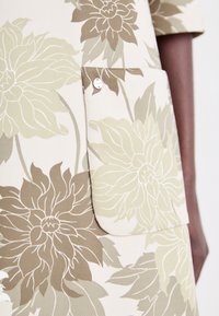 Robe fleurie aux tons beige et vert olive. Présente de grands motifs floraux, une poche poitrine avec un bouton décoratif, et une texture de tissu lisse.