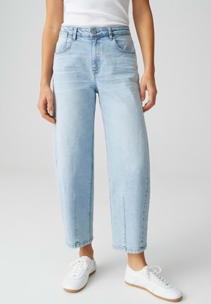 Person trägt hellblaue Jeans mit hohem Bund und geradem Bein, weiße Sneakers und ein weißes ärmelloses Top vor einfarbigem Hintergrund.