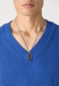 Blå strikket sweater med dyb V-hals, parret med et sølv kædevedhæng med en rektangulær sort pendel.